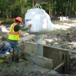 Large machinery holding a bag that is pouring the clay particles of AquaBlok into a contained area.