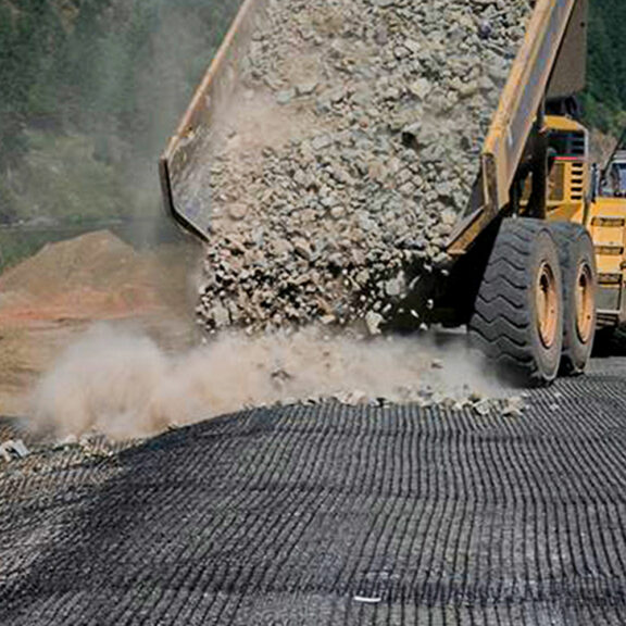 Large dump truck spreading aggregate on top of a geogrid to reduce fill expense and increase stabilization